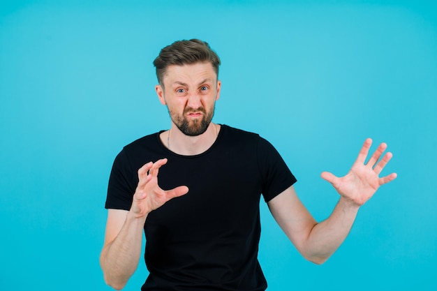 Young man with wild mimicry is showing claws gestures on blue background