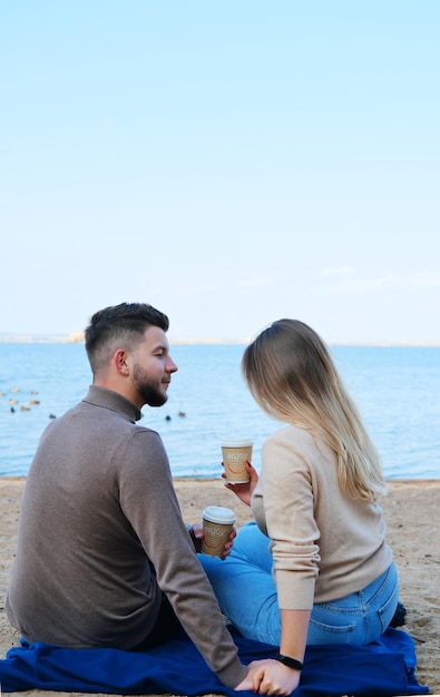 A young man and woman are sitting on the beach with their backs to the camera with glasses of coffee in their hands The guy looks at the girl lovingly