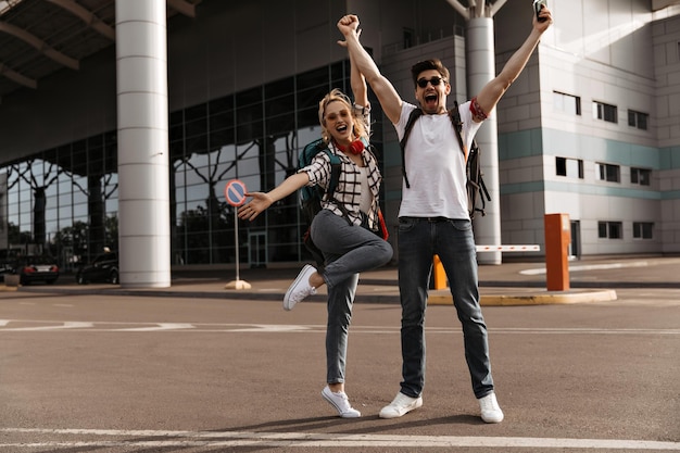 Young man and woman in denim pants and white tees rejoice Couple of travelers with backpacks happily poses near airport