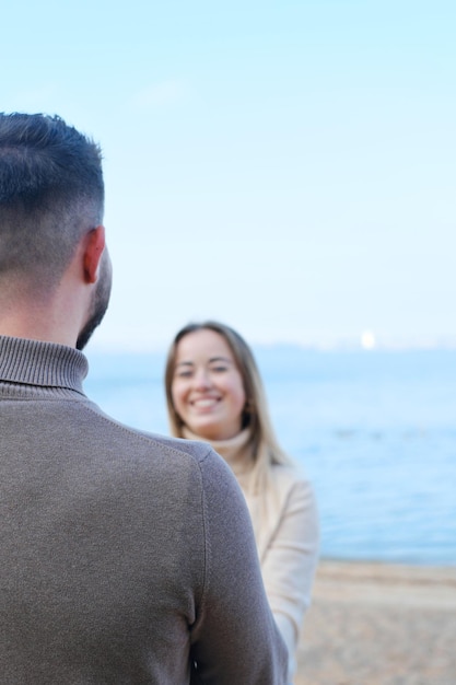 Young man and woman holding hands and posing on the beach against the backdrop of blue water The girl looks at the guy and smiles Focus on the guy's shoulder