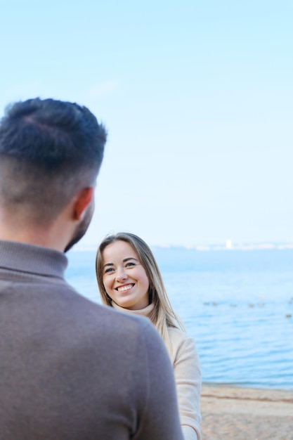 Young man and woman holding hands and posing on the beach against the backdrop of blue water The girl looks at the guy and smiles