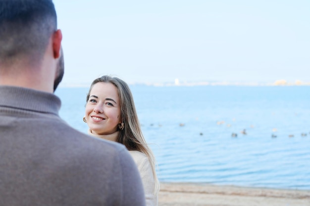 Young man and woman holding hands and posing on the beach against the backdrop of blue water The girl looks at the guy and smiles