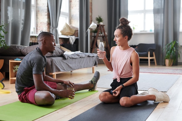 Young man and woman in love sitting on floor in living room at home drinking water and chatting after morning workout