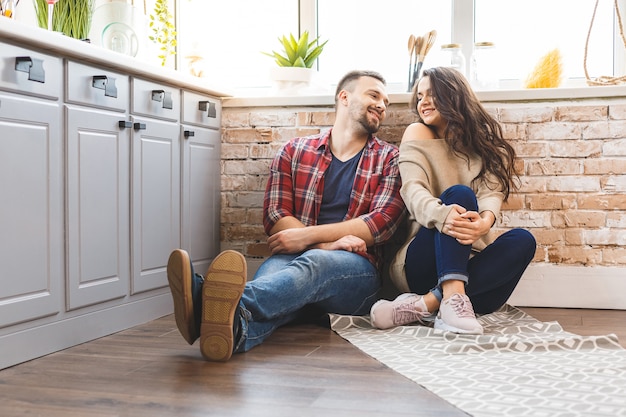 Young man and woman sitting on floor in kitchen and talking. Loving young couple spending time together at home.