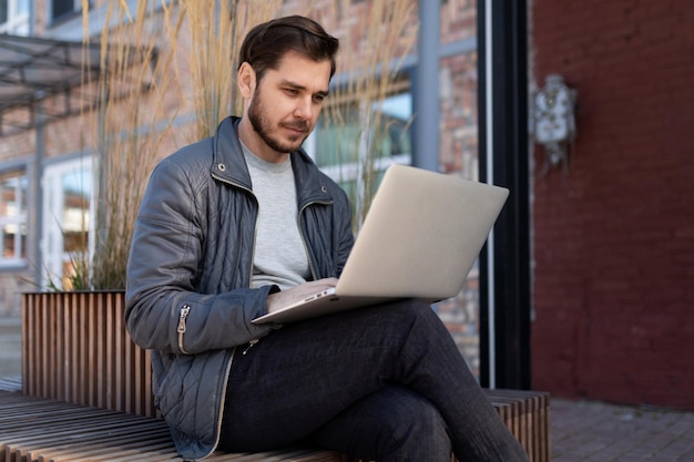 Young man working on a laptop on the street next to a brick building