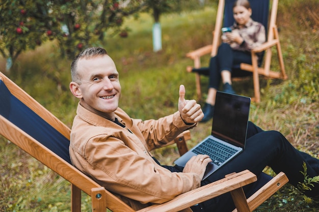 A young man works in the garden outdoors while sitting in a comfortable garden chair Remote work on a portable laptop