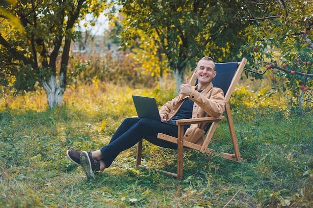 A young man works on a laptop outdoors A young freelancer rests in the forest Remote work active recreation in the summer Tourism people concept man sitting on a chair outdoors