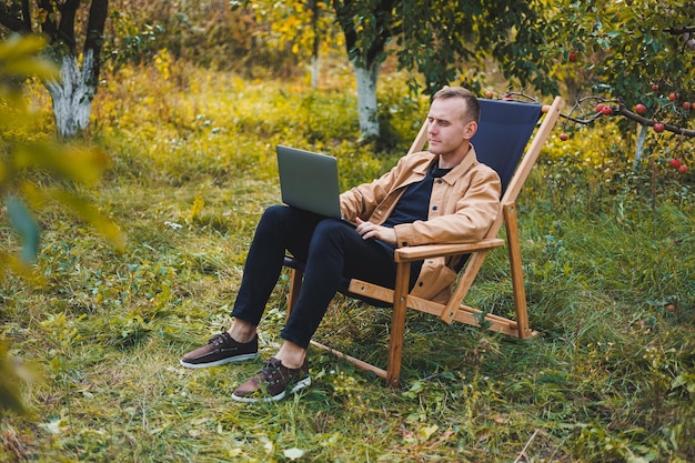 A young man works on a laptop outdoors A young freelancer rests in the forest Remote work active recreation in the summer Tourism people concept man sitting on a chair outdoors