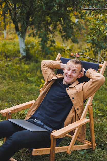 Photo a young man works on a laptop outdoors a young freelancer rests in the forest remote work active recreation in the summer tourism people concept man sitting on a chair outdoors