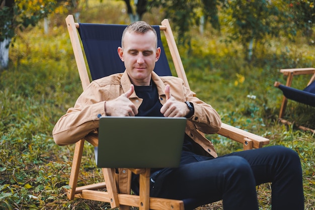 A young man works on a laptop outdoors A young freelancer rests in the forest Remote work active recreation in the summer Tourism people concept man sitting on a chair outdoors