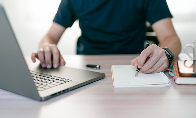 Young man writing down on notebook and using laptop computer sitting at home office