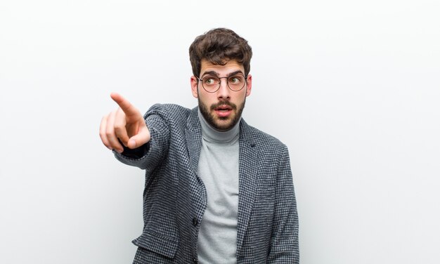 Young manager man feeling shocked and surprised, pointing and looking upwards in awe with amazed, open-mouthed look against white wall