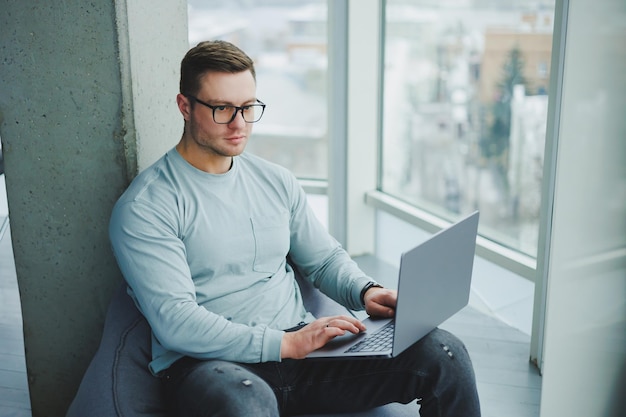 A young manager man in glasses and casual clothes sits on a soft chair and works on a laptop Work space for remote work