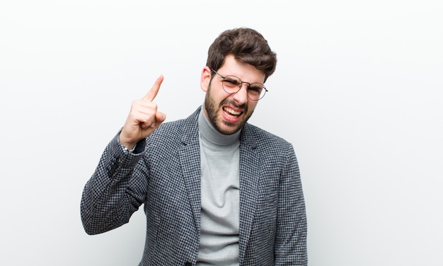 Young manager man pointing with an angry aggressive expression looking like a furious, crazy boss against white wall