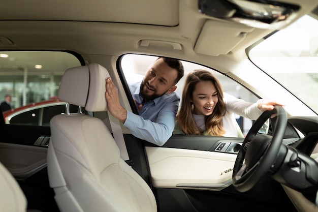 A young married couple examines with interest the features of the interior of a new car in a car dealership