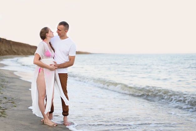 Young married couple on the sea coast. Husband hugs his pregnant wife. Pre-sunset time