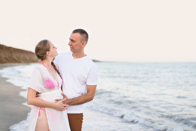 Young married couple on the sea coast. Husband hugs his pregnant wife. Pre-sunset time