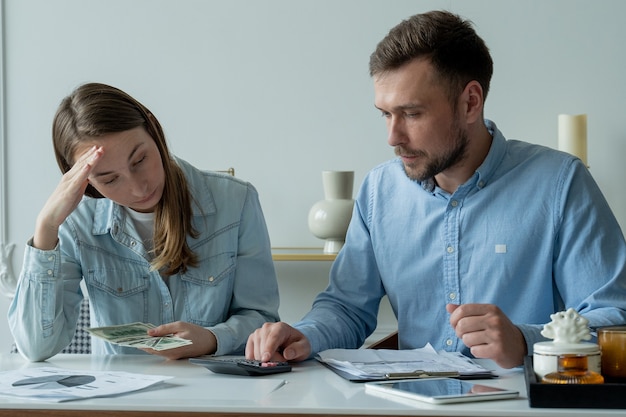 Young married couple sitting at living room table studying papers makes calculations on a calculator studies the family budget