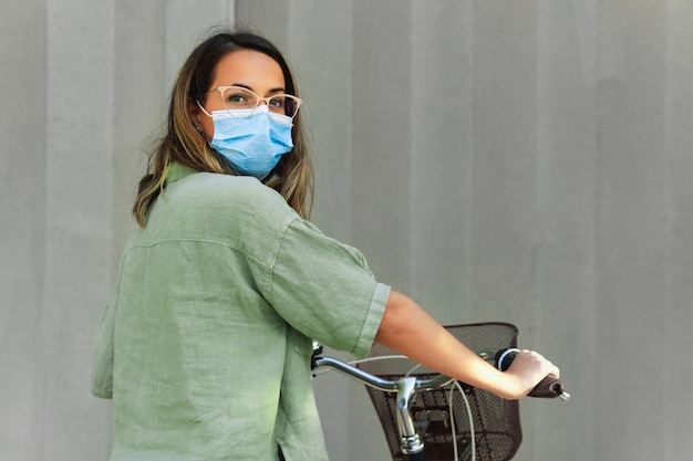 Young masked woman holding a bicycle in a grey background