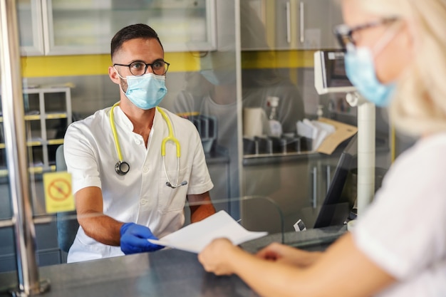 Young medical worker with rubber gloves, facial mask, in sterile uniform giving test results to a patient while sitting in laboratory.