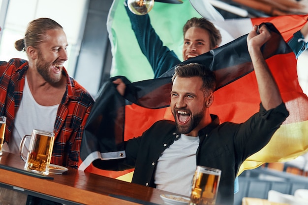 Young men covered in international flags enjoying beer while watching sport game in the pub