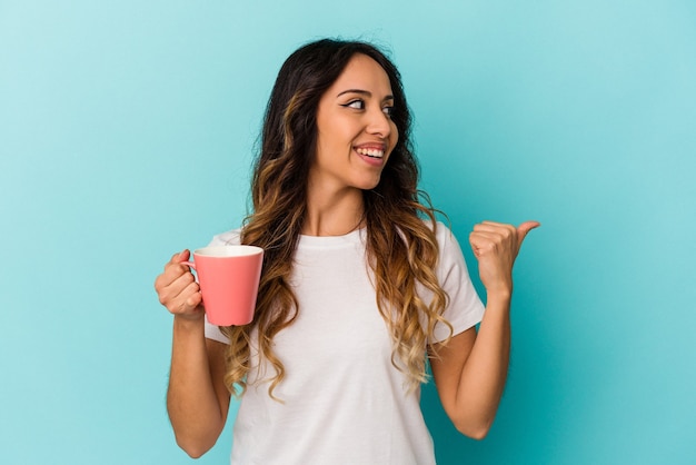 Young mexican woman holding a mug isolated on blue background points with thumb finger away, laughing and carefree.