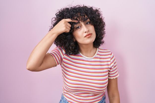 Young middle east woman standing over pink background pointing unhappy to pimple on forehead ugly infection of blackhead acne and skin problem