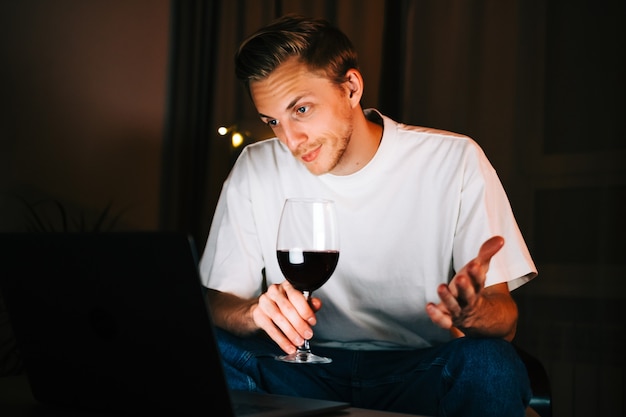 Young millennial man having video call on laptop computer and drinking wine, use technology for communicate with friends or family.