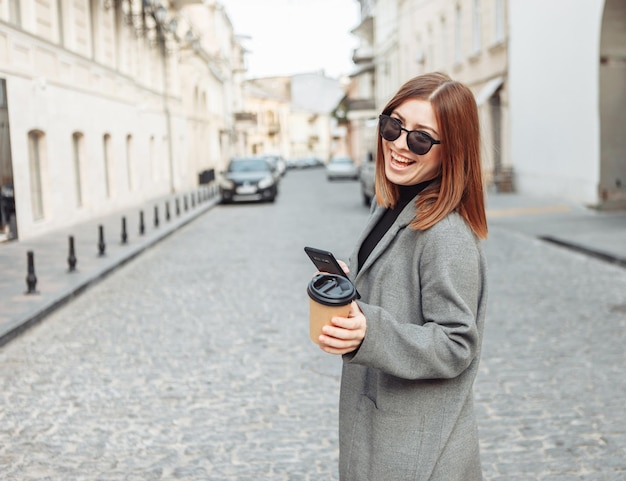 Young millennial smiling woman with smooth hair dressed in an autumn coat and sunglasses uses a smartphone and holds a cup of coffee on the go in a European city.