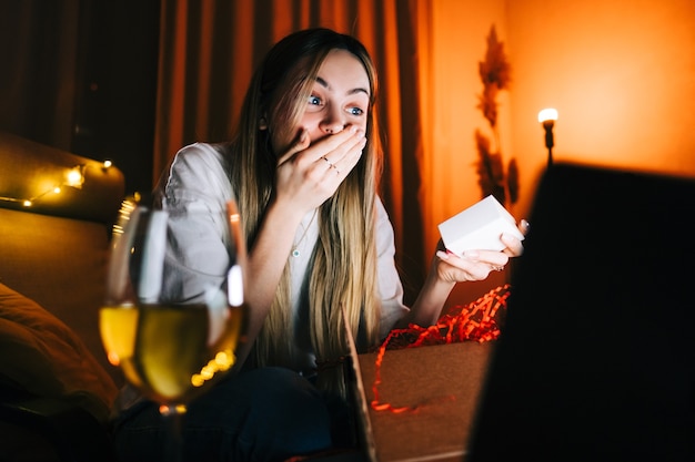 Young millennial woman open gift in front laptop computer during video call and drinking wine, use technology for communicate with friends or family.