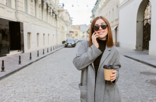 Young millennial woman with smooth hair dressed in autumn coat and sunglasses talking on phone and holds cup of coffee on the go in European city.