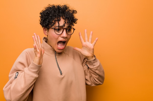 Young mixed african american woman against a brown wall shouts loud, keeps eyes opened and hands tense.