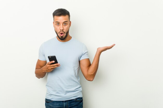 Young mixed race asian man holding a phone impressed holding blank space on palm.