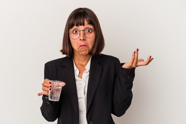 Young mixed race business woman holding a glass of water isolated on white background shrugs shoulders and open eyes confused.