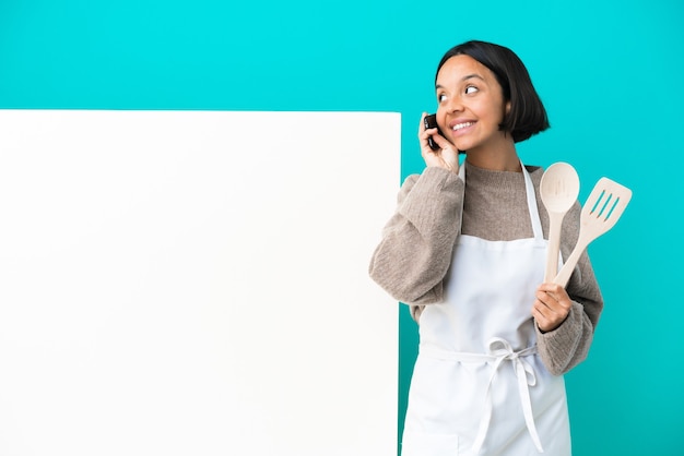 Young mixed race cook woman with a big placard isolated on blue wall keeping a conversation with the mobile phone with someone