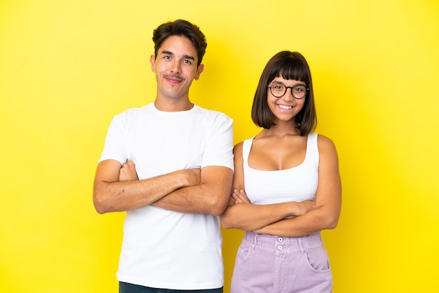 Young mixed race couple isolated on yellow background keeping the arms crossed in frontal position