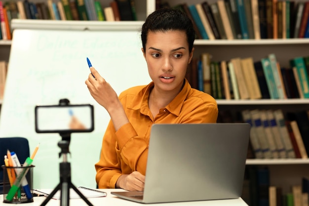 Young mixed race female teacher in glasses talking to laptop webcam and explaining new material to students