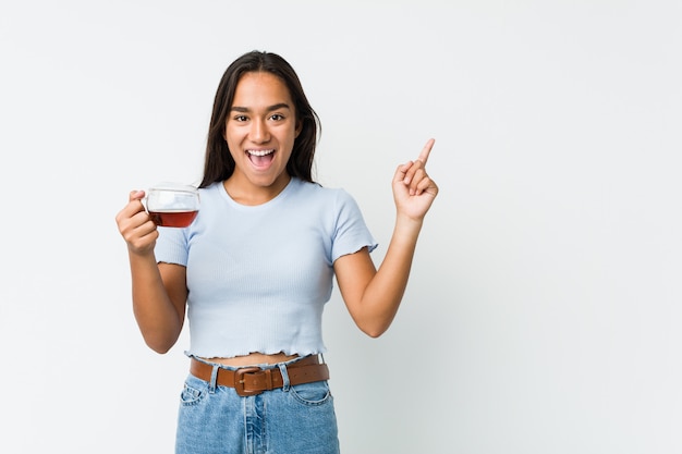 Young mixed race indian holding a tea cup smiling cheerfully pointing with forefinger away.