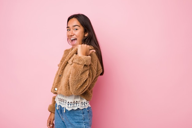 Young mixed race indian woman wearing a short sheepskin coatpoints with thumb finger away, laughing and carefree.