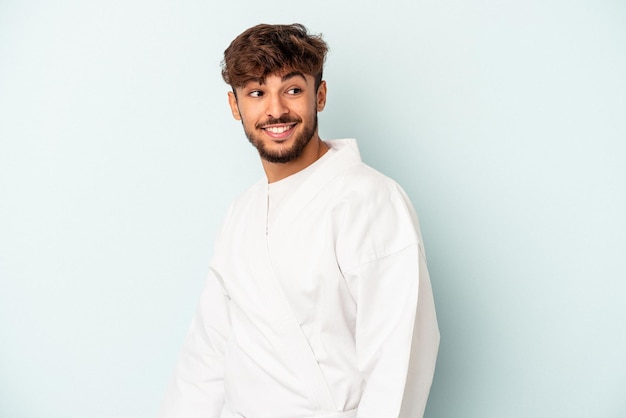 Young mixed race man doing karate isolated on blue background looks aside smiling, cheerful and pleasant.