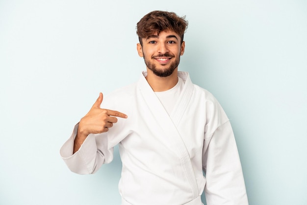 Young mixed race man doing karate isolated on blue background person pointing by hand to a shirt copy space, proud and confident