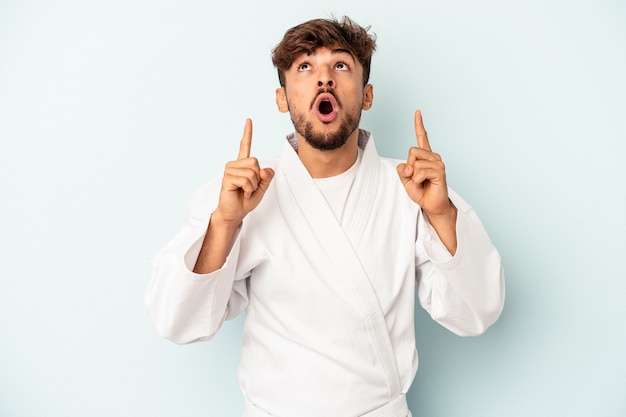 Young mixed race man doing karate isolated on blue background pointing upside with opened mouth.