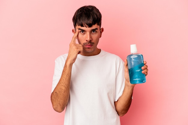 Young mixed race man holding mouthwash isolated on pink background pointing temple with finger, thinking, focused on a task.