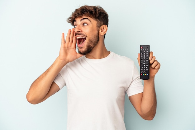 Young mixed race man holding a tv controller isolated on blue background shouting and holding palm near opened mouth.