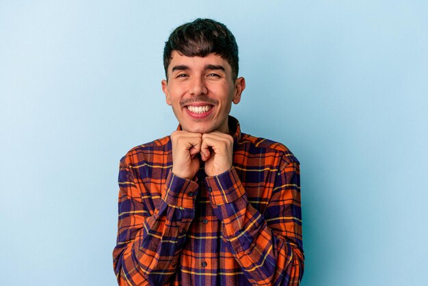 Young mixed race man isolated on blue background keeps hands under chin, is looking happily aside.