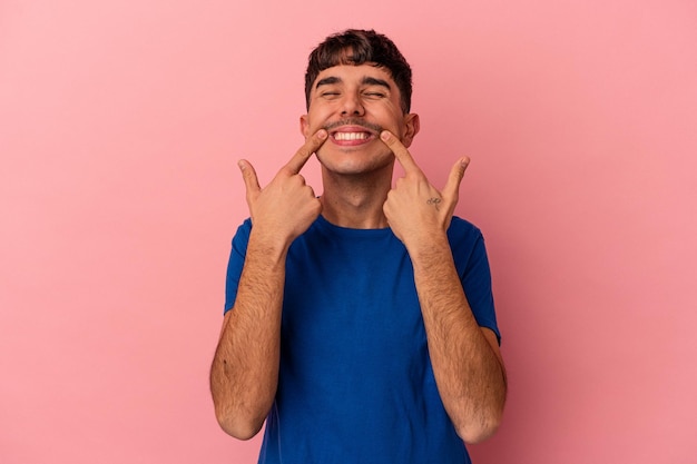 Young mixed race man isolated on pink background smiles pointing fingers at mouth