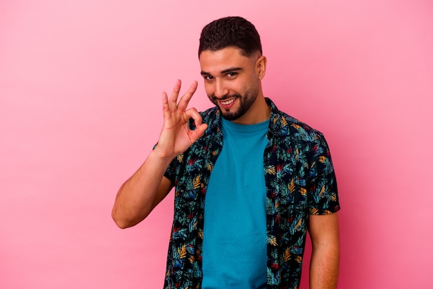 Young mixed race man isolated on pink background winks an eye and holds an okay gesture with hand.