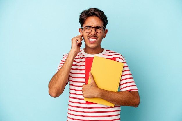 Young mixed race student man holding books isolated on blue background covering ears with hands.