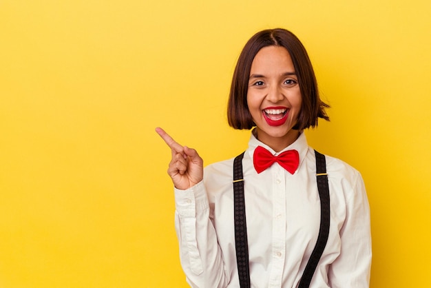 Young mixed race waitress woman isolated on yellow background smiling and pointing aside, showing something at blank space.