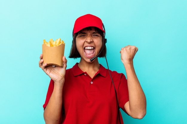 Young mixed race woman fast food restaurant worker holding fries isolated on blue background raising fist after a victory, winner concept.